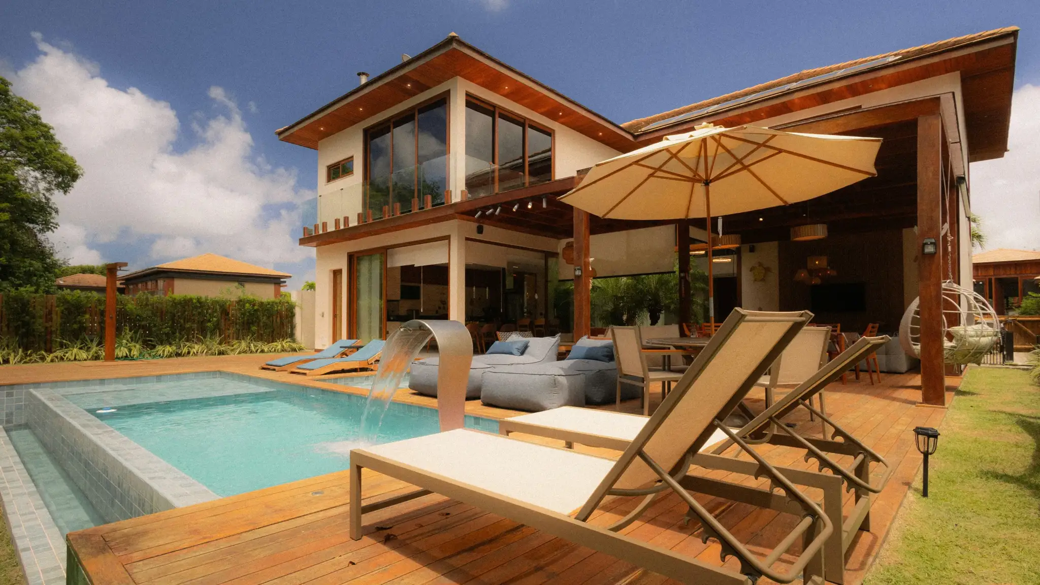 Piscină din beton într-o curte amenajată, cu șezlonguri, umbrelă și cascadă decorativă