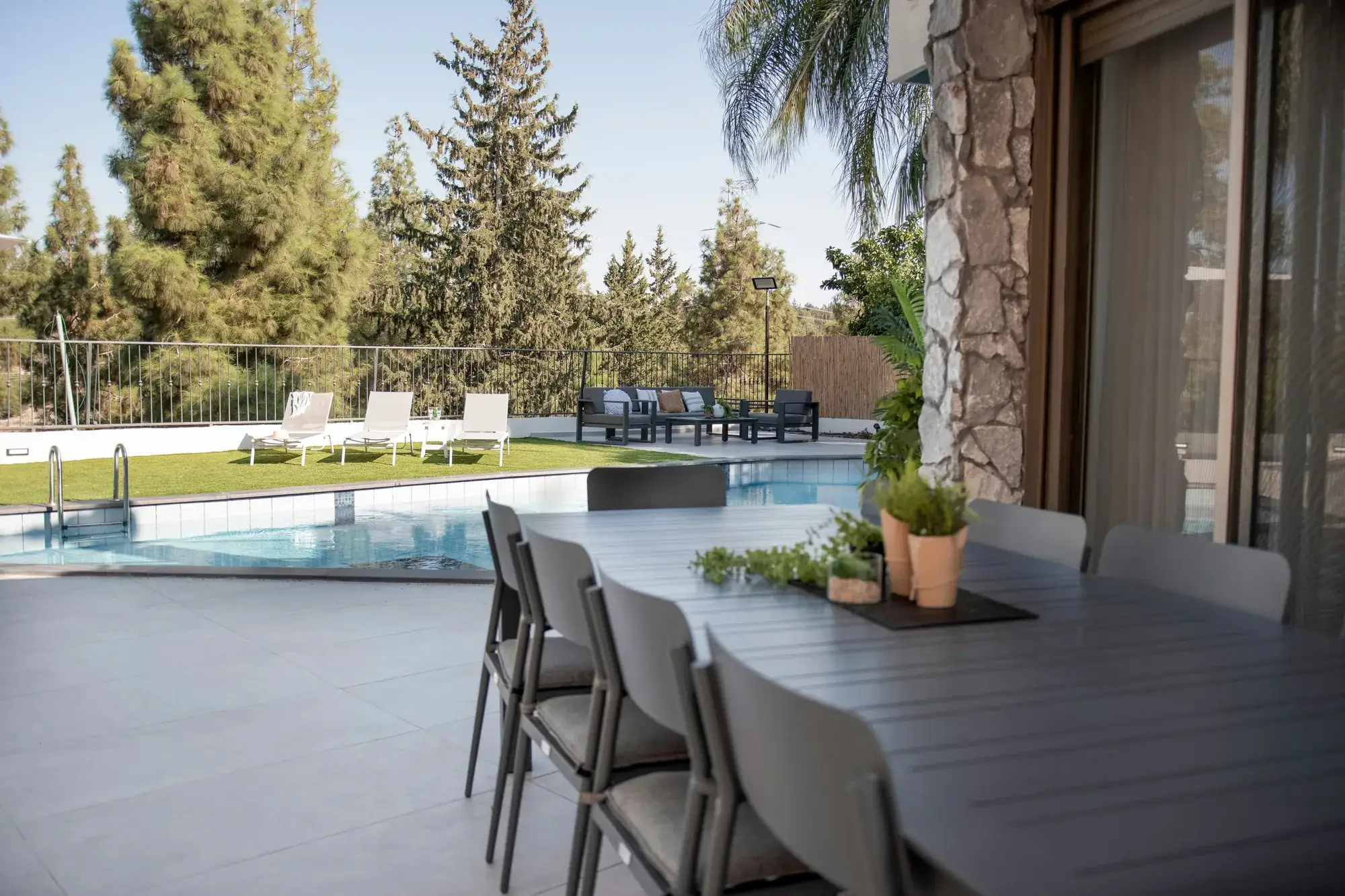 Piscină văzută de pe terasa acoperită, cu masă de exterior în prim-plan și șezlonguri pe gazon
